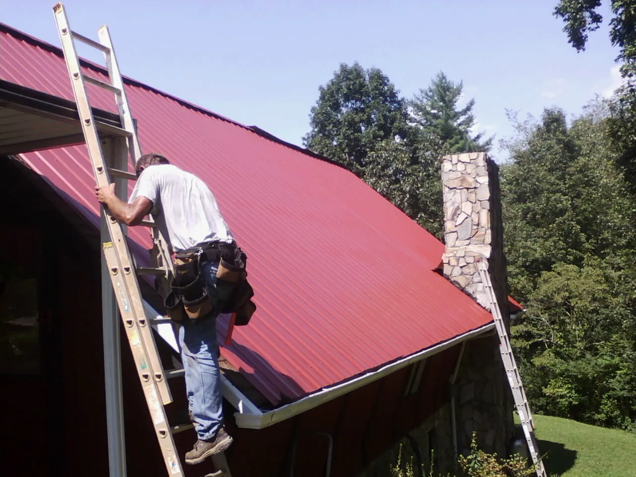 Roofer on a ladder beside a red metal roof and stone chimney — Soffit & Fascia Repair by Roofing Brothers in Plainfield